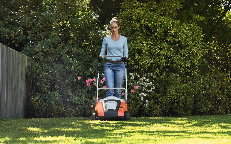 Woman with a lawn scarifier on the lawn of a garden with flowering bushes in the background and a wooden fence, frontal